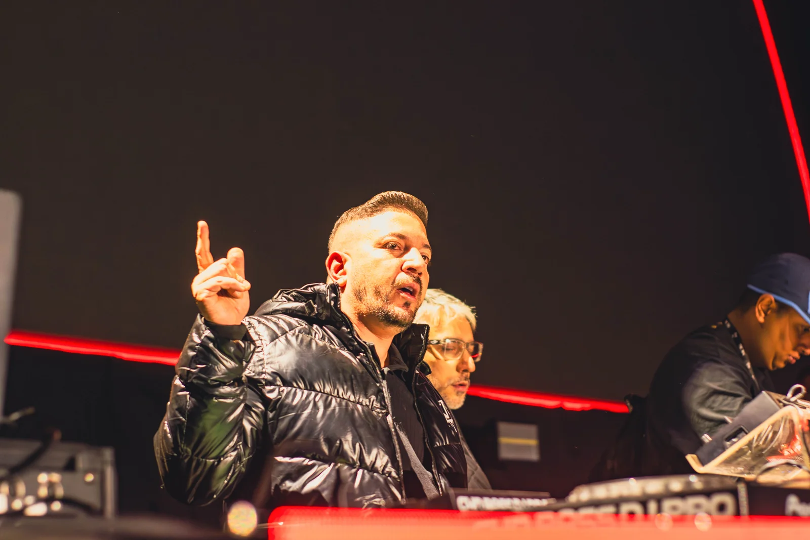 Godoy em apresentação diante dos equipamentos de DJ, com jaqueta escura e iluminação vermelha.