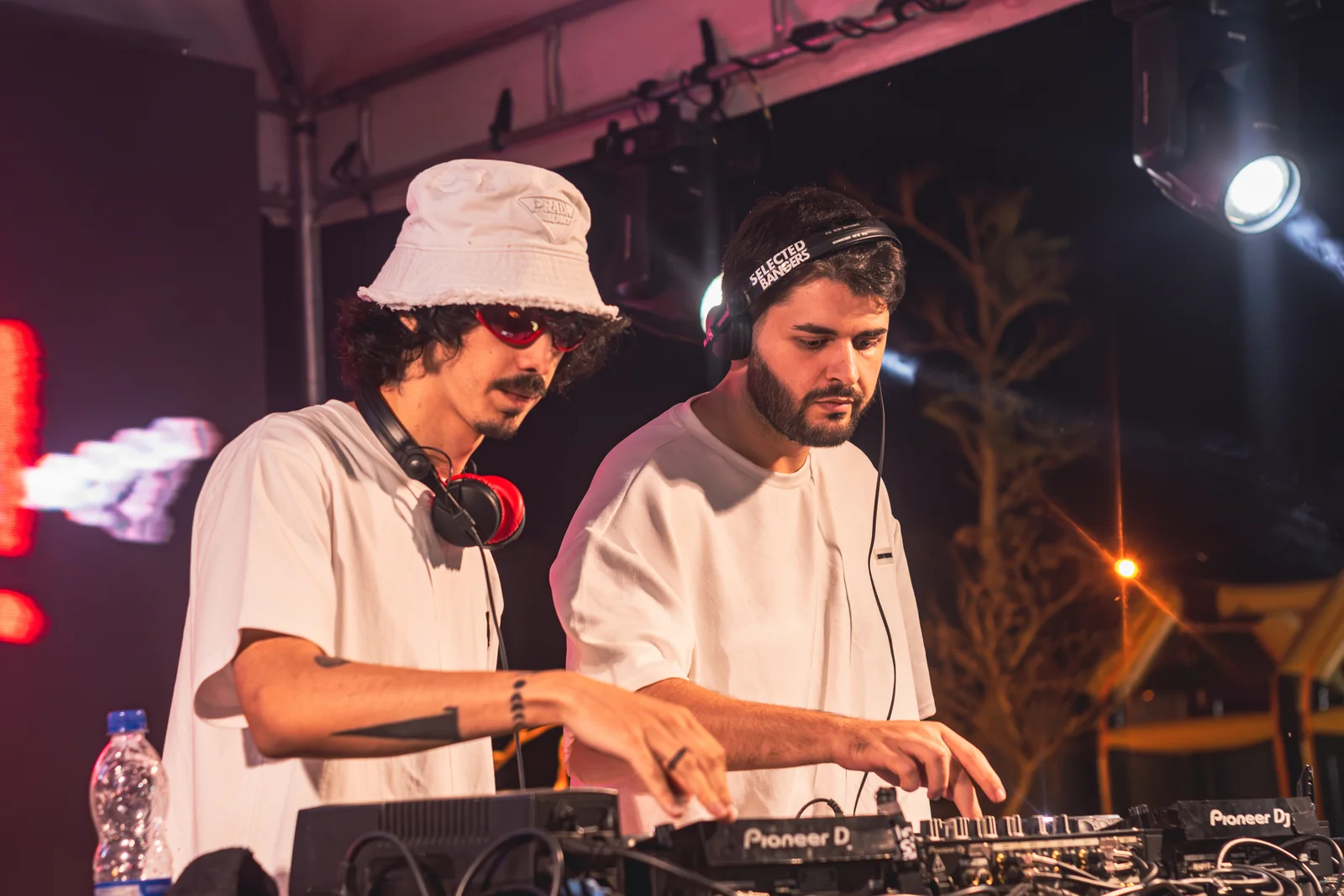 HeyDoc em dupla diante dos equipamentos de DJ, com iluminação de palco.